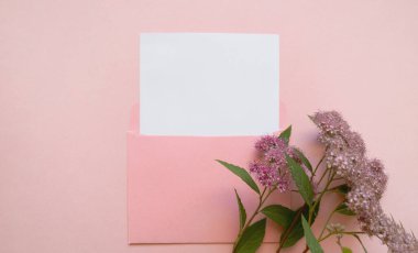 Pembe bir zarf, yanında beyaz bir maket kağıdı ve pembe bir çiçek var..