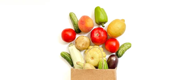 Una bolsa de compras de papel de la que salen verduras y frutas, a saber, tomate, pepino, calabaza, pimienta, limón, berenjena, calabacín, plátano, manzana, melocotón sobre fondo blanco. — Foto de Stock