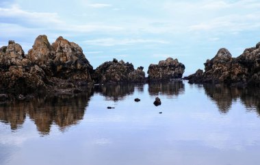 Sakin ve durgun sudan yükselen engebeli kahverengi kayaların olduğu sakin bir kıyı manzarası. Denizin pürüzsüz, ayna benzeri yüzeyi oluşumları ve yumuşak, bulutlu mavi gökyüzünü mükemmel bir şekilde yansıtır.
