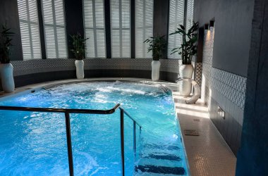 Kapalı yüzme havuzu ve köpüren mavi suyu olan bir spa. Seramikli duvarlar, beyaz kepenkler, yeşil bitkiler ve rahatlama için hidroterapi ekipmanlarıyla modern sağlık merkezi..