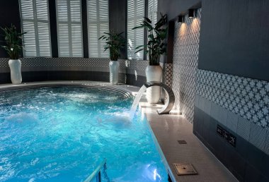 Kapalı yüzme havuzu ve köpüren mavi suyu olan bir spa. Seramikli duvarlar, beyaz kepenkler, yeşil bitkiler ve rahatlama için hidroterapi ekipmanlarıyla modern sağlık merkezi..