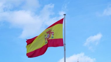 İspanya 'nın ulusal bayrağının alçak açılı görüntüsü rüzgarda gururla dalgalanan beyaz bulutlu açık mavi gökyüzüne karşı. Parlak kırmızı ve sarı renkler armalı gök mavisi karşısında duruyor.