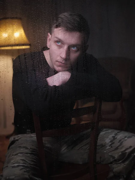 A man in camouflage pants sits by a rainy window