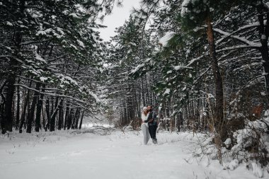 Bir çift, karlı bir ormanda, arka plandaki ağaçlara sarılıyor.