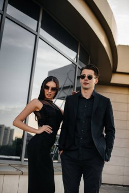 Siyah elbiseli ve gözlüklü güzel, şık bir çift genç gün batımında bir ofis binasının arka planında duruyor. Moda ve tarz