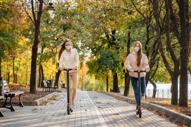 Maskeli iki genç kız sıcak bir sonbahar gününde parkta elektrikli scooterlara biniyor. Parkta yürü..