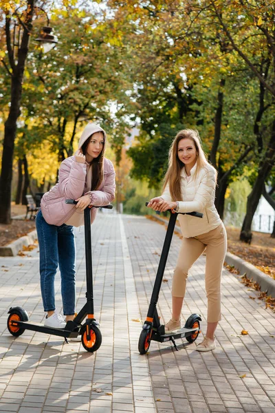 2人の若い美しい女の子は暖かい秋の日に公園で電動スクーターに乗る — ストック写真