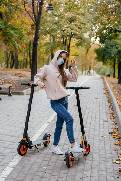 マスクの若い美しい女の子が暖かい秋の日に電動スクーターで公園に乗っています 公園を歩く — ストック写真