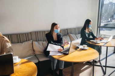 Bir grup maskeli kız dizüstü bilgisayarlarla çalışırken bir kafede sosyal mesafelerini koruyorlar..
