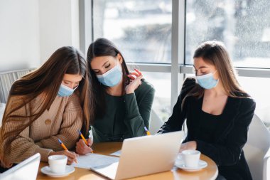 Maskeli bir grup kız bir kafede oturup dizüstü bilgisayarlarla çalışıyor. Öğrenciler.