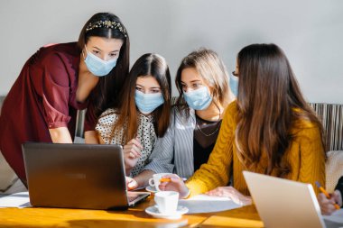 Maskeli bir grup kız bir kafede oturup dizüstü bilgisayarlarla çalışıyor. Öğrenciler.