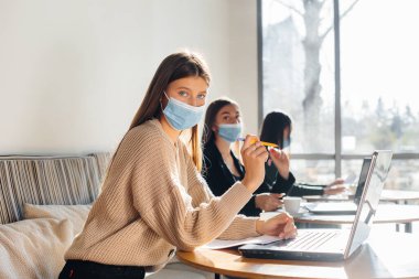 Bir grup maskeli kız dizüstü bilgisayarlarla çalışırken bir kafede sosyal mesafelerini koruyorlar..
