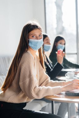 Bir grup maskeli kız dizüstü bilgisayarlarla çalışırken bir kafede sosyal mesafelerini koruyorlar..