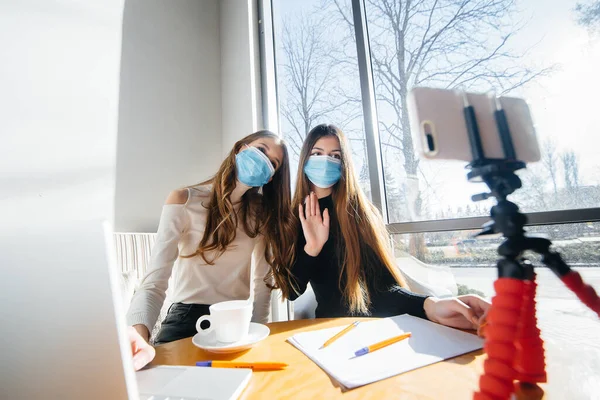 Two young girls sit in a cafe in masks and lead a video blog. Communication to the camera.