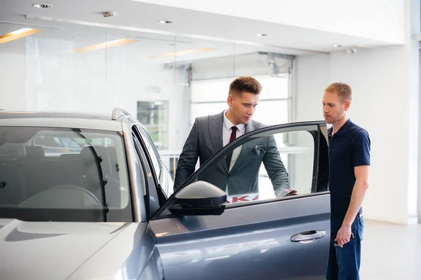 一个年轻的商人和一个推销员一起看汽车经销店里的一辆新车 买一辆汽车 — 图库照片