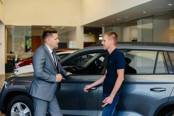 一个年轻的商人和一个推销员一起看汽车经销店里的一辆新车 买一辆汽车 — 图库照片