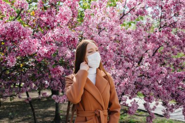 Genç bir kız maskesini çıkarıyor ve salgından sonra derin derin nefes alıyor güneşli bir bahar gününde, çiçek açan bahçelerin önünde. Koruma ve önleme bölmesi 19