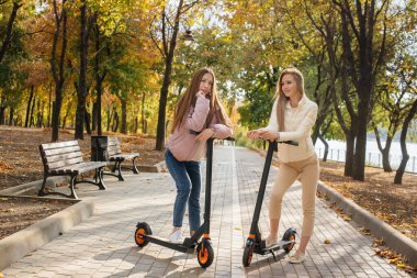 Ilık bir sonbahar gününde iki güzel kız parkta elektrikli scooterlara biniyor.