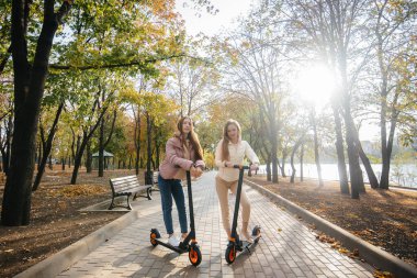 Ilık bir sonbahar gününde iki güzel kız parkta elektrikli scooterlara biniyor.