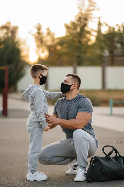 A father and child stand on a sports field in masks after training during sunset