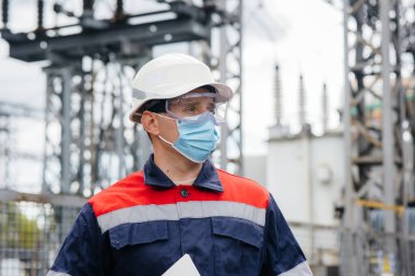 Bir elektrik santrali mühendisi Pondemia zamanında maske takan modern yüksek voltajlı ekipmanları inceliyor. Enerji. Sanayi