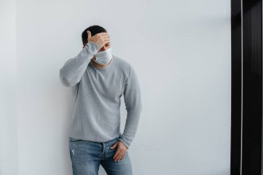 Genç bir adam, karantina sırasında maske takarak gri arka planda duruyor. Maskeli karantina