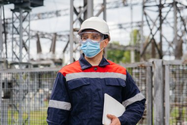 Bir elektrik santrali mühendisi Pondemia zamanında maske takan modern yüksek voltajlı ekipmanları inceliyor. Enerji. Sanayi