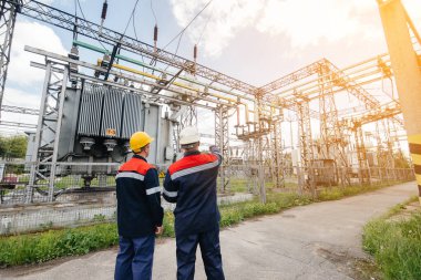 İki uzman elektrik santrali mühendisi günbatımında modern yüksek voltajlı ekipmanları inceliyor. Enerji. Sanayi