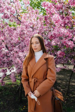 Genç bir kız maskesini çıkarıyor ve salgından sonra derin derin nefes alıyor güneşli bir bahar gününde, çiçek açan bahçelerin önünde. Koruma ve önleme bölmesi 19