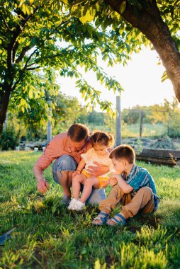 Küçük erkek ve kız kardeşler gün batımında babalarıyla güzel bir bahar bahçesinde fidanlar dikiyorlar. Yeni bir hayat. Çevreyi koru. Çevredeki dünyaya ve doğaya karşı dikkatli olun.