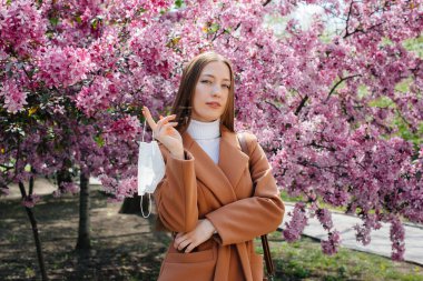 Genç bir kız maskesini çıkarıyor ve salgından sonra derin derin nefes alıyor güneşli bir bahar gününde, çiçek açan bahçelerin önünde. Koruma ve önleme bölmesi 19