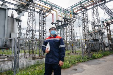 Bir elektrik santrali mühendisi Pondemia zamanında maske takan modern yüksek voltajlı ekipmanları inceliyor. Enerji. Sanayi