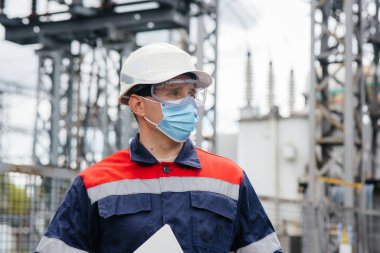 Bir elektrik santrali mühendisi Pondemia zamanında maske takan modern yüksek voltajlı ekipmanları inceliyor. Enerji. Sanayi