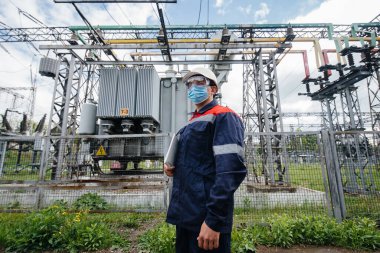 Bir elektrik santrali mühendisi Pondemia zamanında maske takan modern yüksek voltajlı ekipmanları inceliyor. Enerji. Sanayi