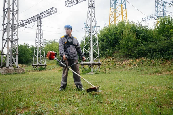 A young man mowing grass on the territory of an electric substation in overalls. Grass cleaning at the enterprise, implementation of fire safety measures