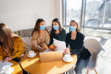 Maskeli bir grup kız bir kafede oturup dizüstü bilgisayarlarla çalışıyor. Öğrenciler.