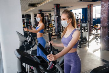 Salgın sırasında maske takan iki güzel kız spor salonunda egzersiz yapıyor. Halka açık yerlerde sosyal mesafeler.