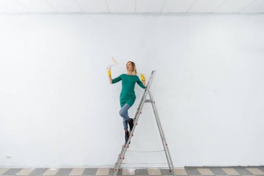 Merdivendeki genç bir kız beyaz bir duvarı silindirle boyuyor. İç kısmın tamiri.