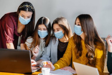 Maskeli bir grup kız bir kafede oturup dizüstü bilgisayarlarla çalışıyor. Öğrenciler.