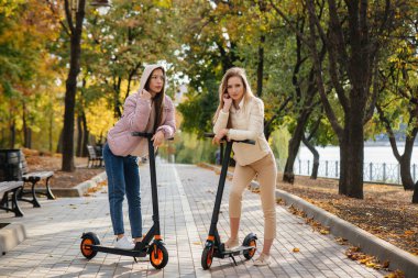 Ilık bir sonbahar gününde iki güzel kız parkta elektrikli scooterlara biniyor.