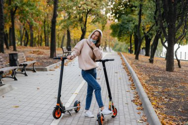 Maskeli genç bir kız sıcak bir sonbahar gününde elektrikli scooterla parkta geziyor. Parkta yürü..