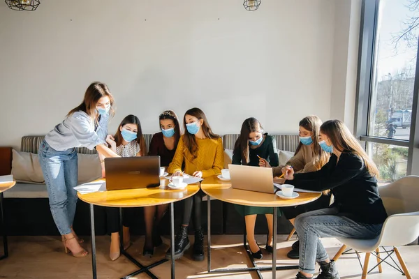 A group of girls in masks sit in a cafe and work on laptops. Teaching students.