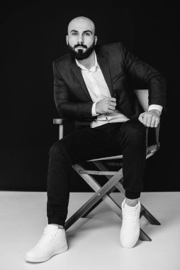 Beyaz gömlekli ve takım elbiseli genç sakallı bir adam siyah arka planda oturuyor. Siyah ve beyaz.