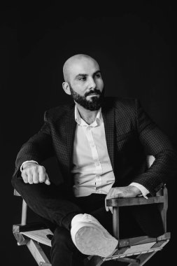Beyaz gömlekli ve takım elbiseli genç sakallı bir adam siyah arka planda oturuyor. Siyah ve beyaz.