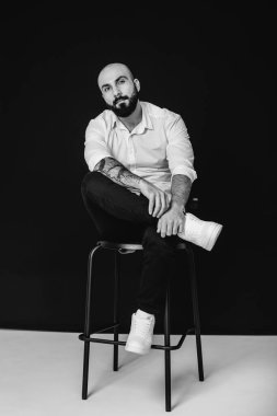 Beyaz gömlekli genç sakallı bir adam siyah arka planda oturuyor. Siyah ve beyaz.