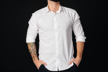 Beyaz gömlekli ve dövmeli genç sakallı bir adam siyah arka planda duruyor..