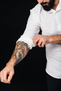Beyaz gömlekli ve dövmeli genç sakallı bir adam siyah arka planda duruyor..