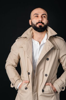 Genç sakallı bir adam dikilir ve siyah bir arka plana karşı yağmurluğunu düzeltir..