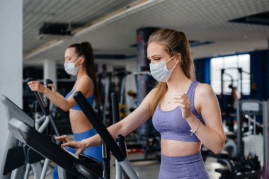 Salgın sırasında maske takan iki güzel kız spor salonunda egzersiz yapıyor. Halka açık yerlerde sosyal mesafeler.