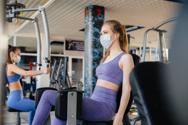 Salgın sırasında maske takan iki güzel kız spor salonunda egzersiz yapıyor. Halka açık yerlerde sosyal mesafeler.